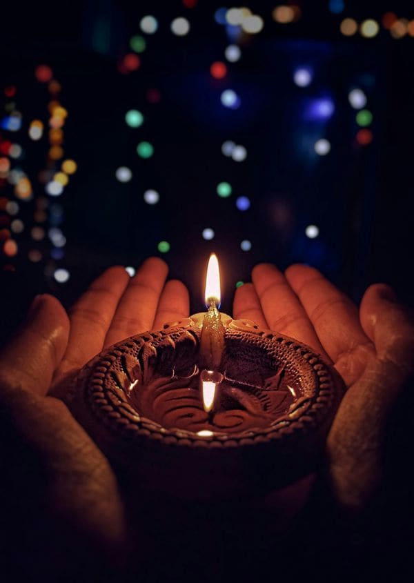 Cupped hands holding a lit clay lamp against festive lights